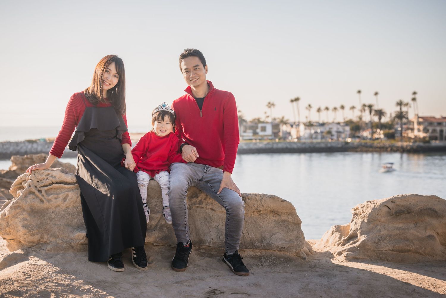 Three people sit on coastal rocks at sunset with palm trees and a marina visible in the background.