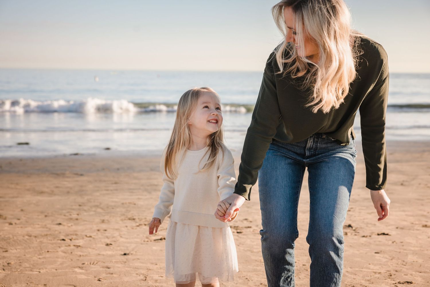 Joyful parent and child holding hands while walking together on a sunny beach near the ocean at sunset.