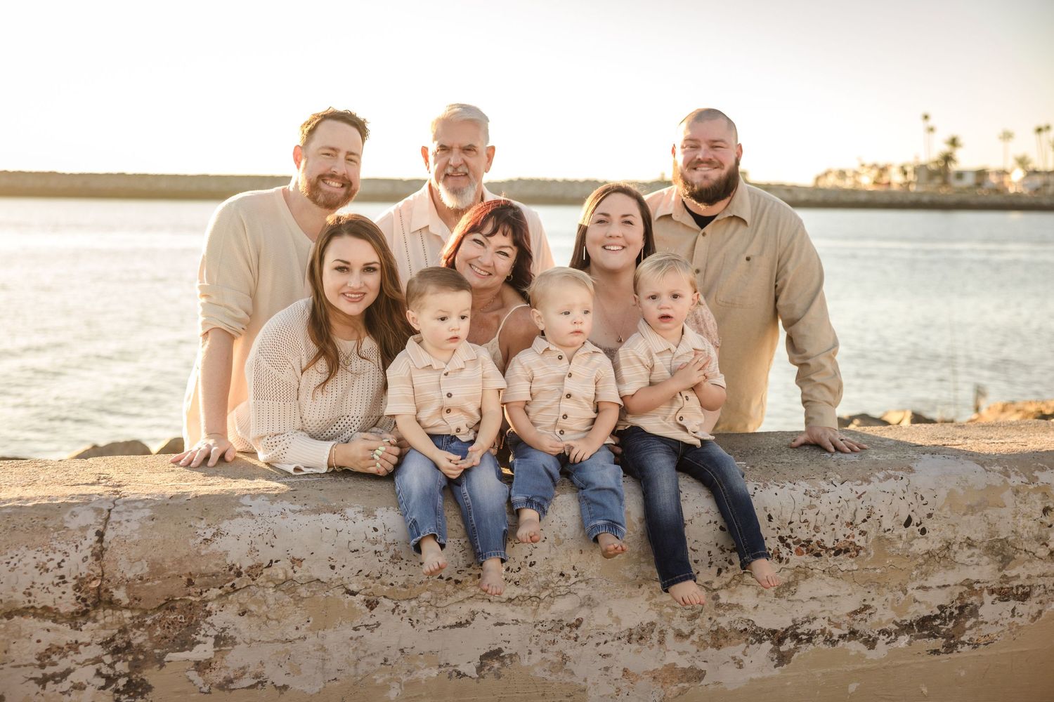 A family group sits together on a rocky beach shore at sunset with tranquil water and landscape in the background.