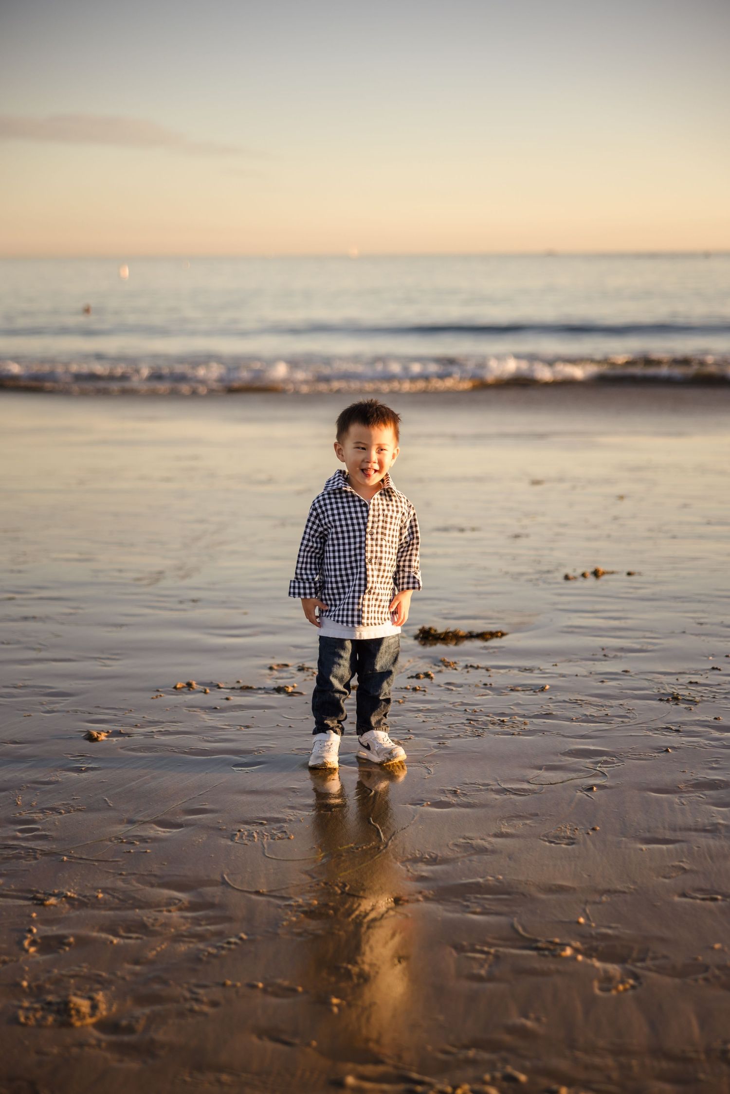 A child stands on a beach at sunset wearing a plaid shirt and boots, with waves and golden light in the background.