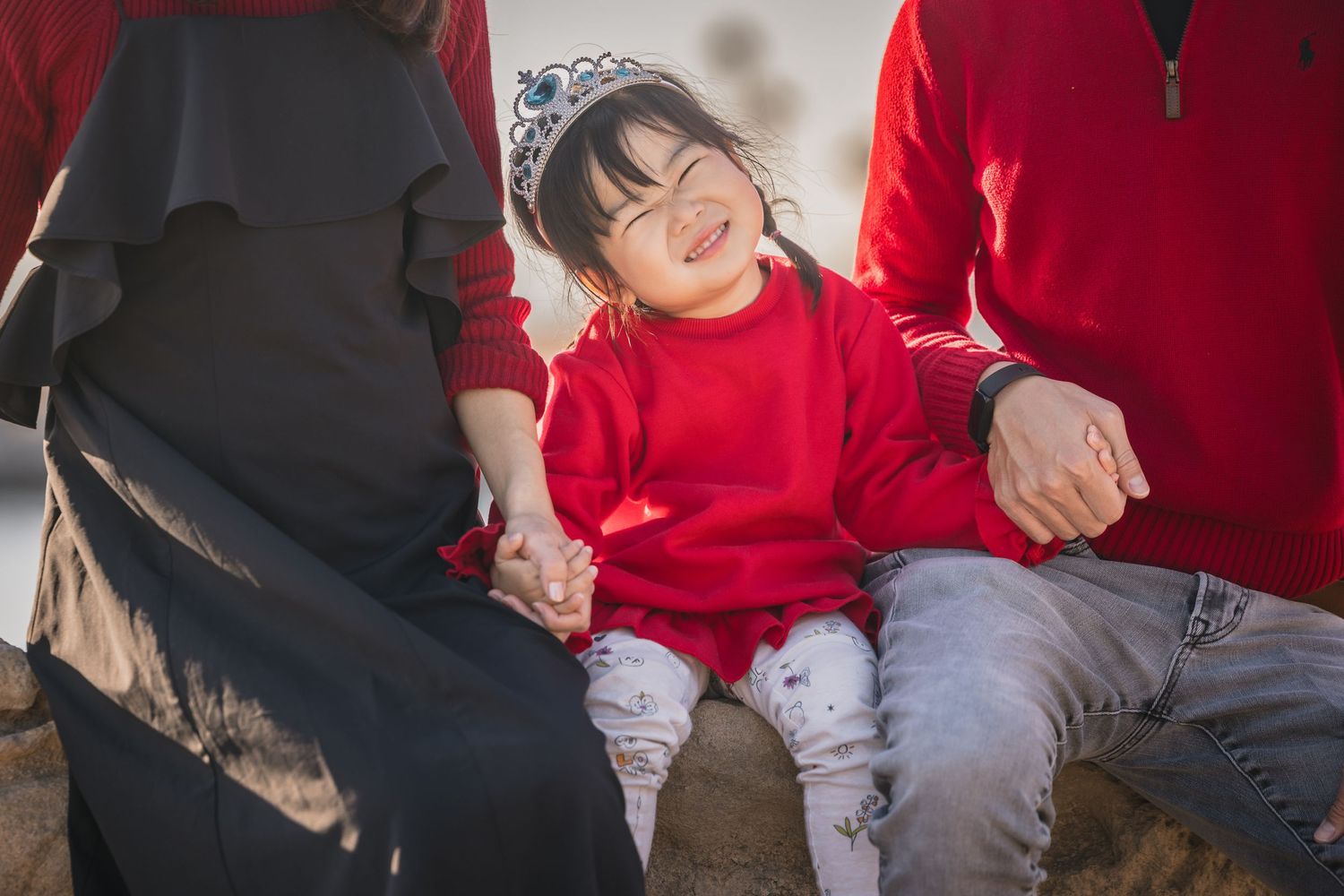 A child wearing a bright red sweater sits outdoors with two adults on a sunny day.