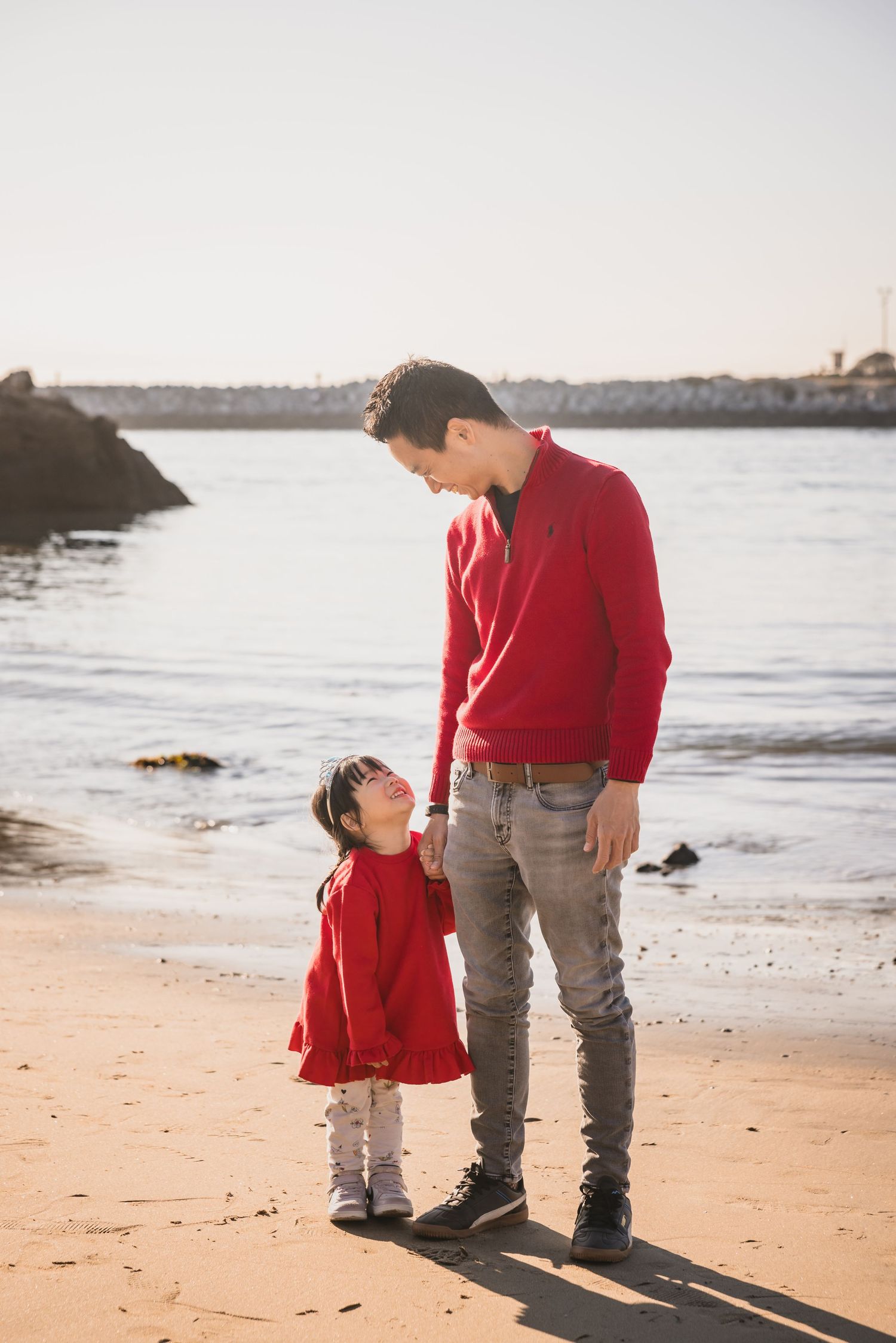 A parent and child in matching red sweaters stand together on a beach at sunset gazing at each other.
