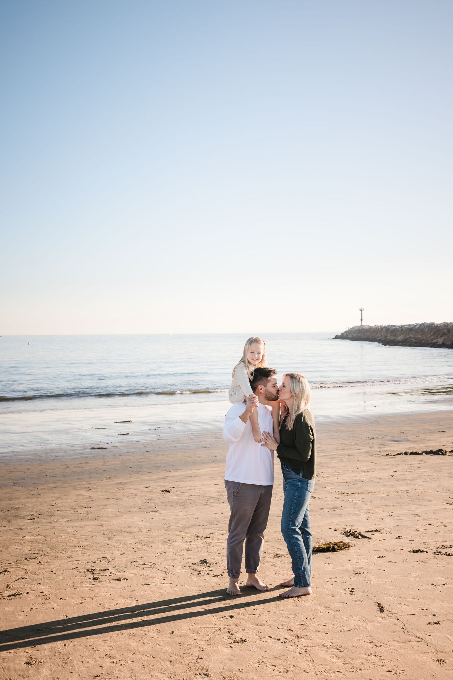 Family enjoying a sunset moment together on a sandy beach with ocean and lighthouse in background.