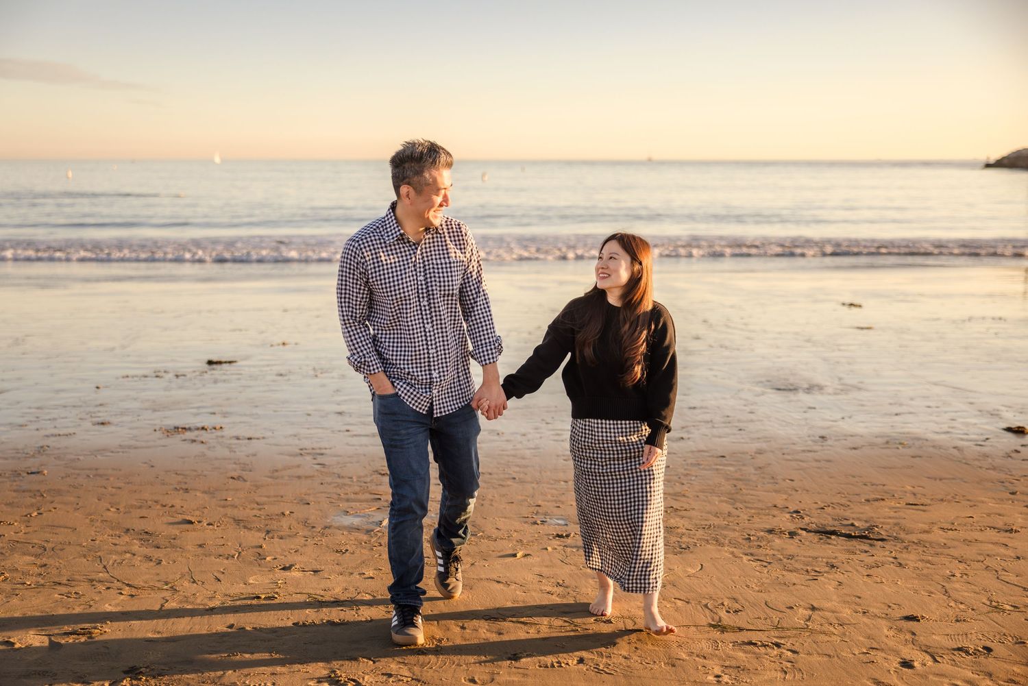 A couple walks hand in hand along a peaceful beach at sunset with golden light illuminating the sandy shoreline.