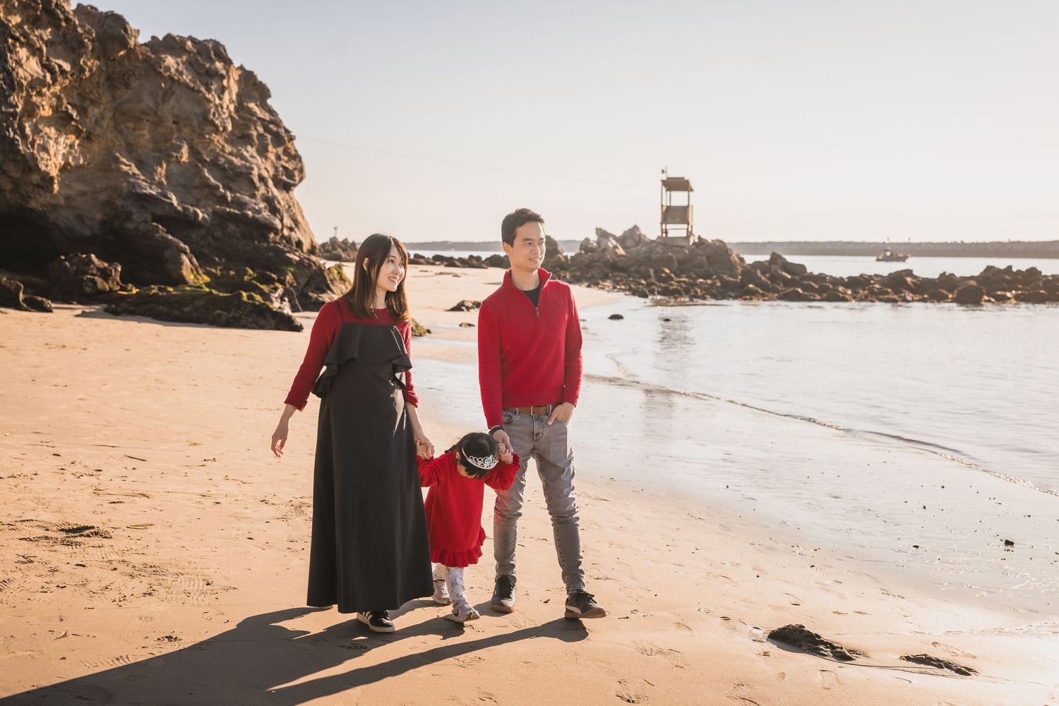A family walks together on a sandy beach with a lighthouse and rocky cliffs in the background on a sunny day.