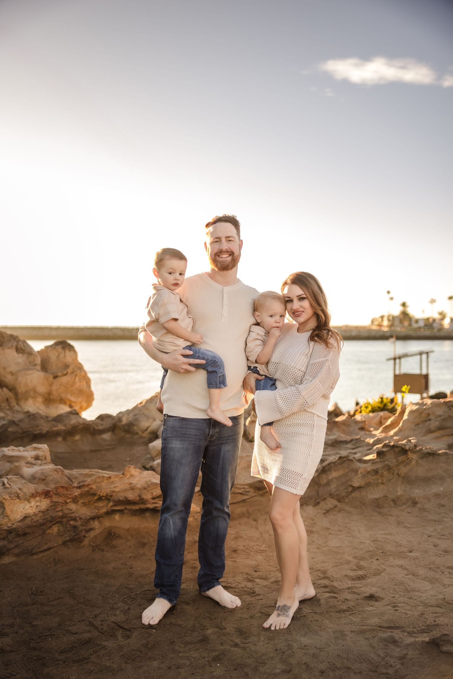 A family of four poses on a beach at sunset with a lighthouse visible in the distance.