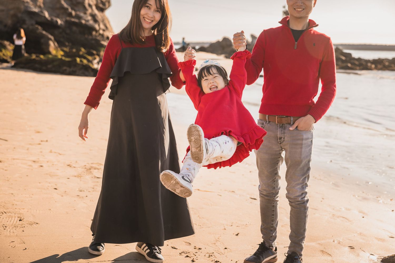 Family wearing red sweaters plays together while swinging child at sunset on a sandy beach.