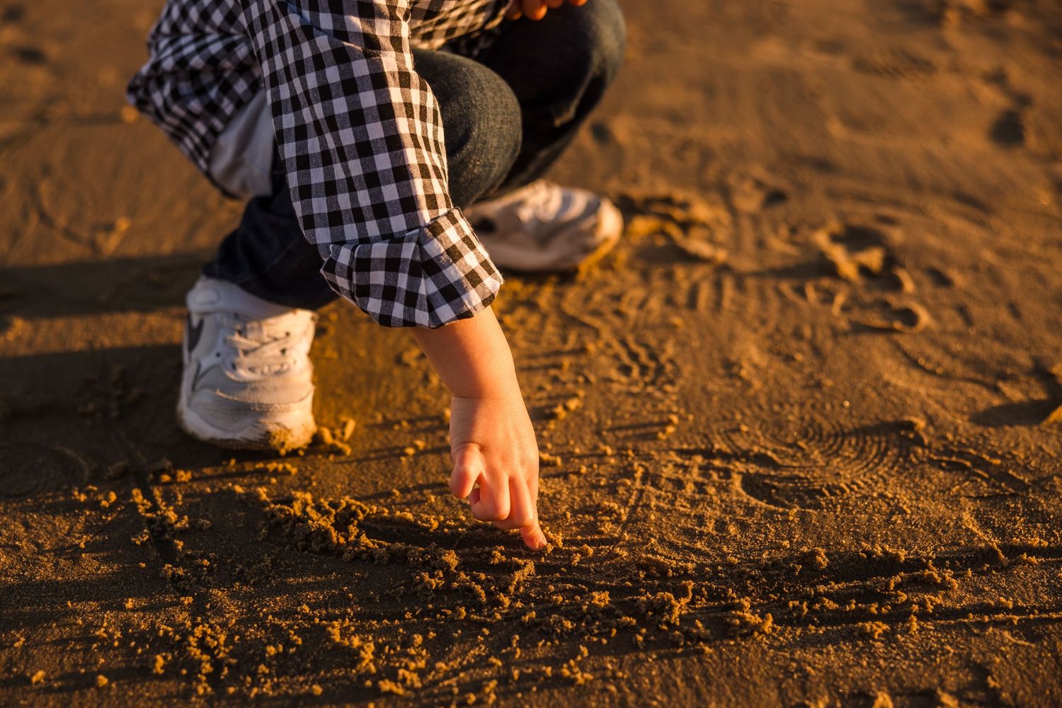 A hand draws in the golden sand at sunset while wearing a plaid shirt and white sneakers.