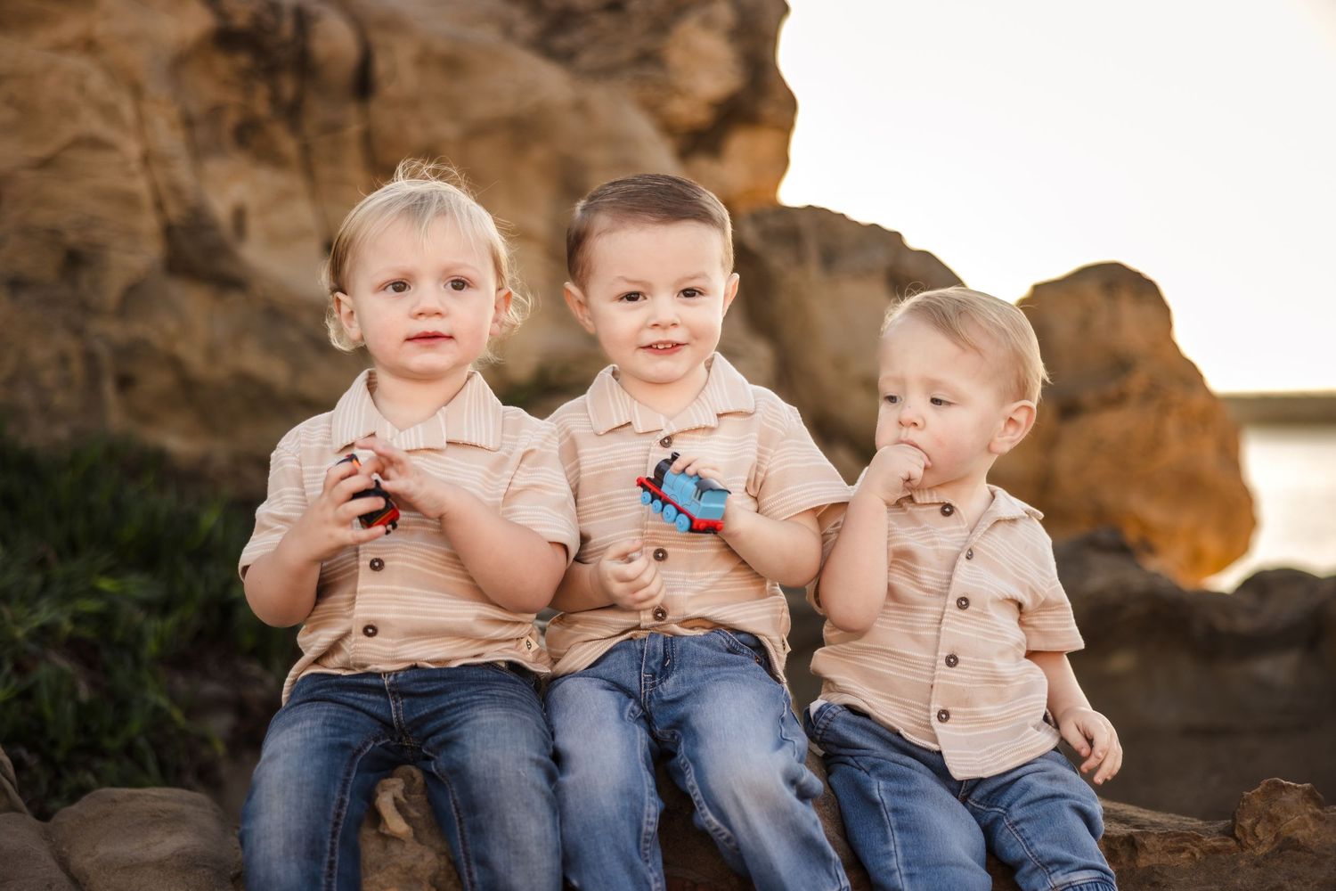 Three children sit together on rocky terrain wearing coordinated neutral-toned outfits at sunset.