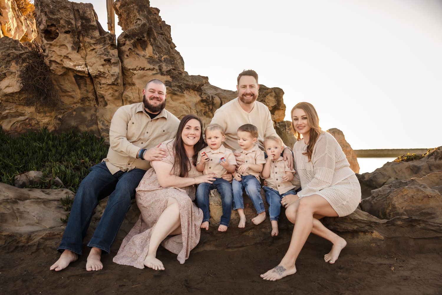 A family group poses together on a rocky beach setting at sunset wearing neutral-colored coordinating outfits.
