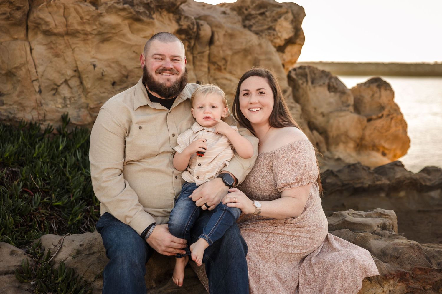 A happy family in casual attire sits together on coastal rocks during a golden hour photoshoot by the ocean.