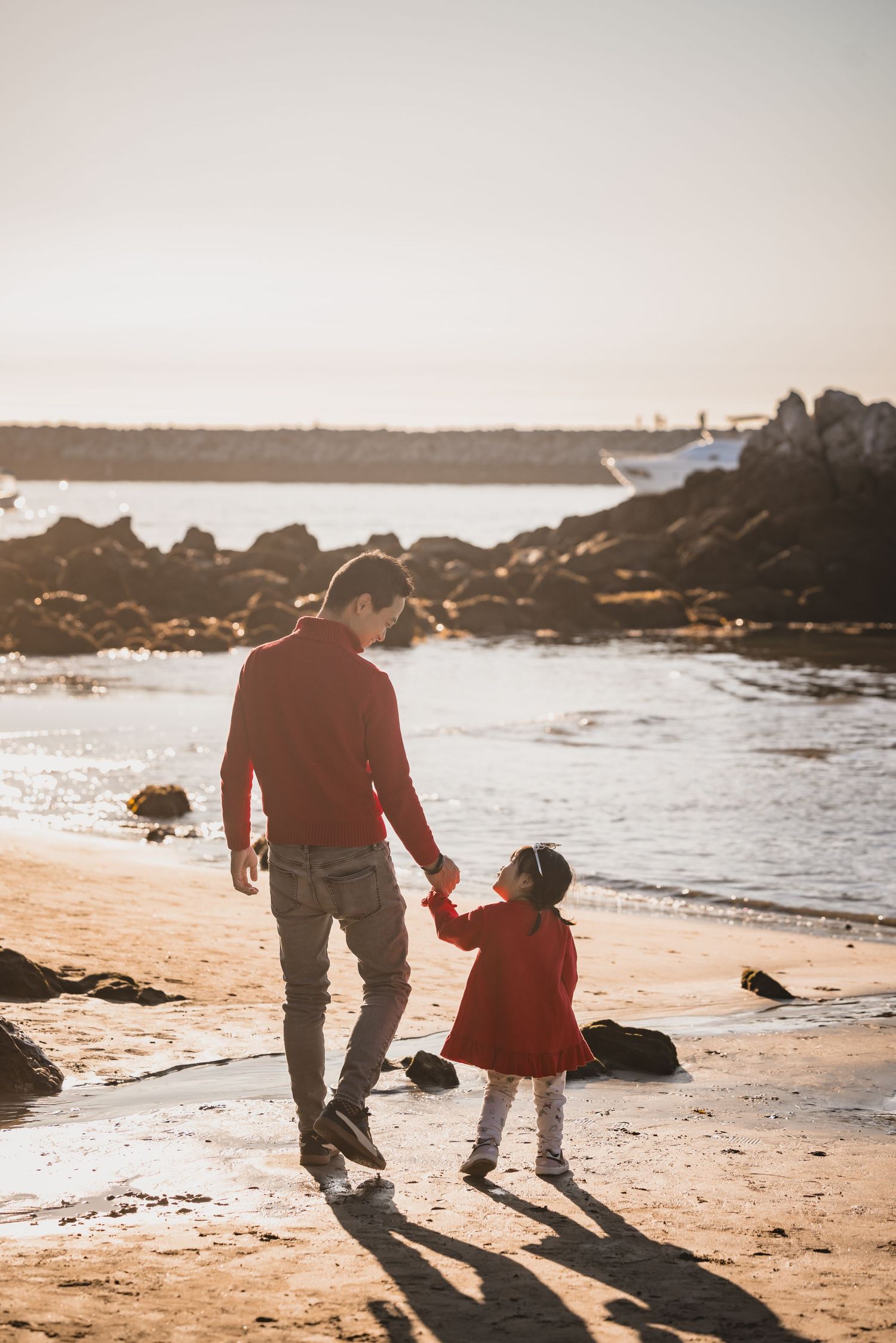 A silhouetted pair walk hand in hand along a rocky beach at sunset as their shadows stretch across the golden sand.