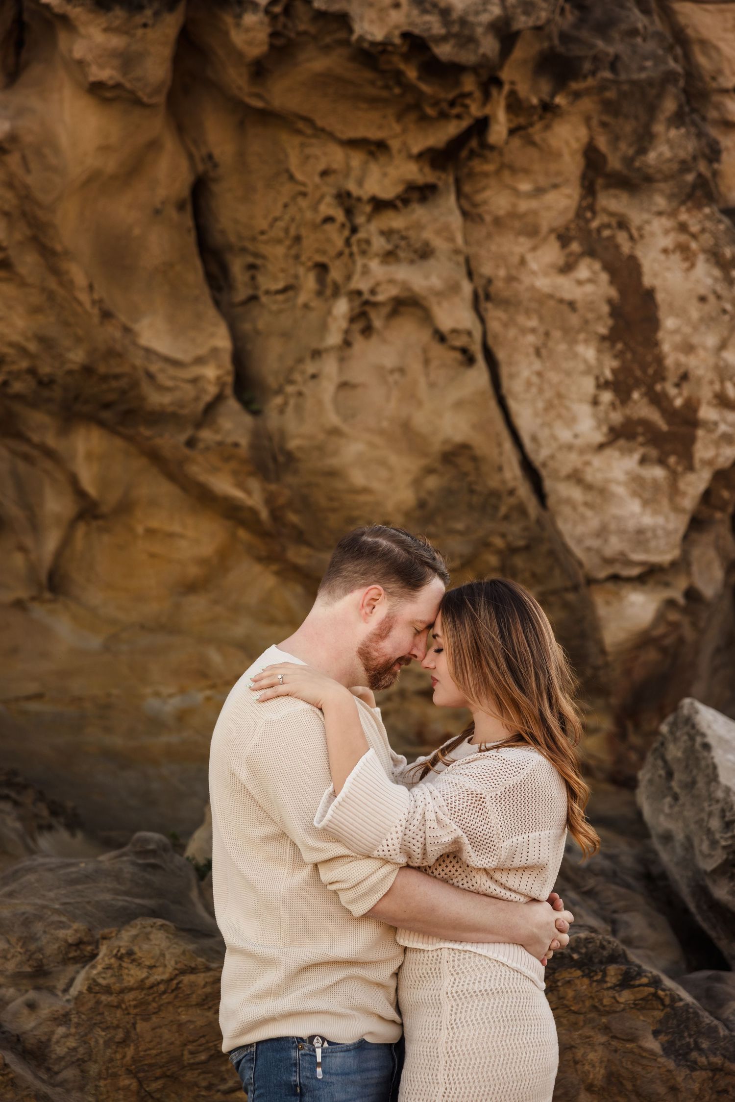 A couple embraces tenderly against a rocky cliff backdrop during a romantic photoshoot at sunset.