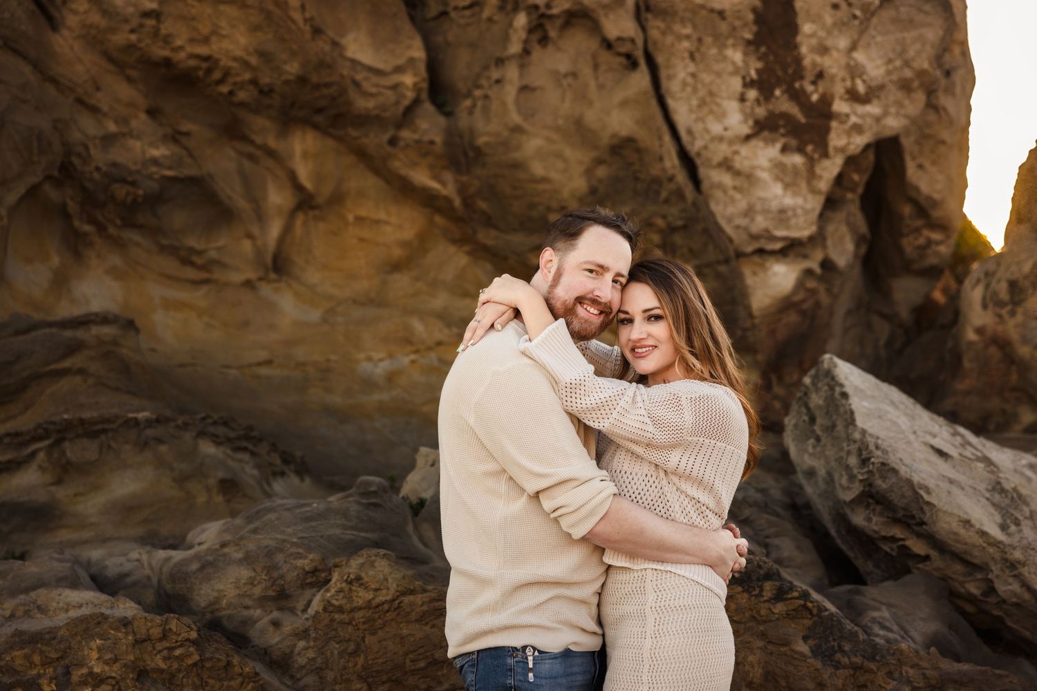 A romantic couple embraces while standing among dramatic rock formations at sunset on the beach.