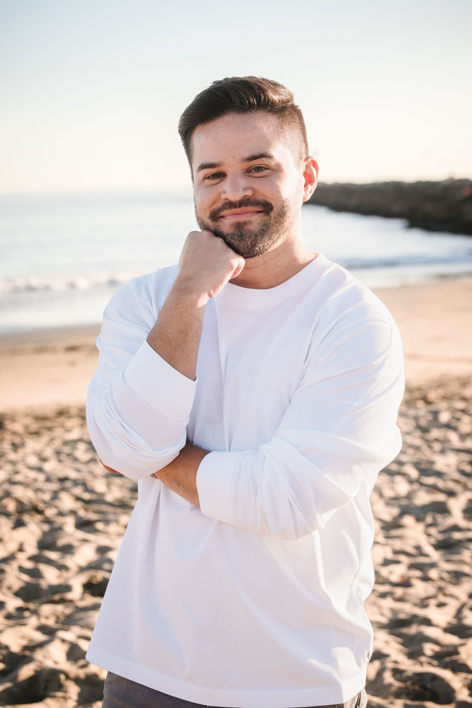 Person in white shirt smiling on beach with scenic coastal background at sunset.