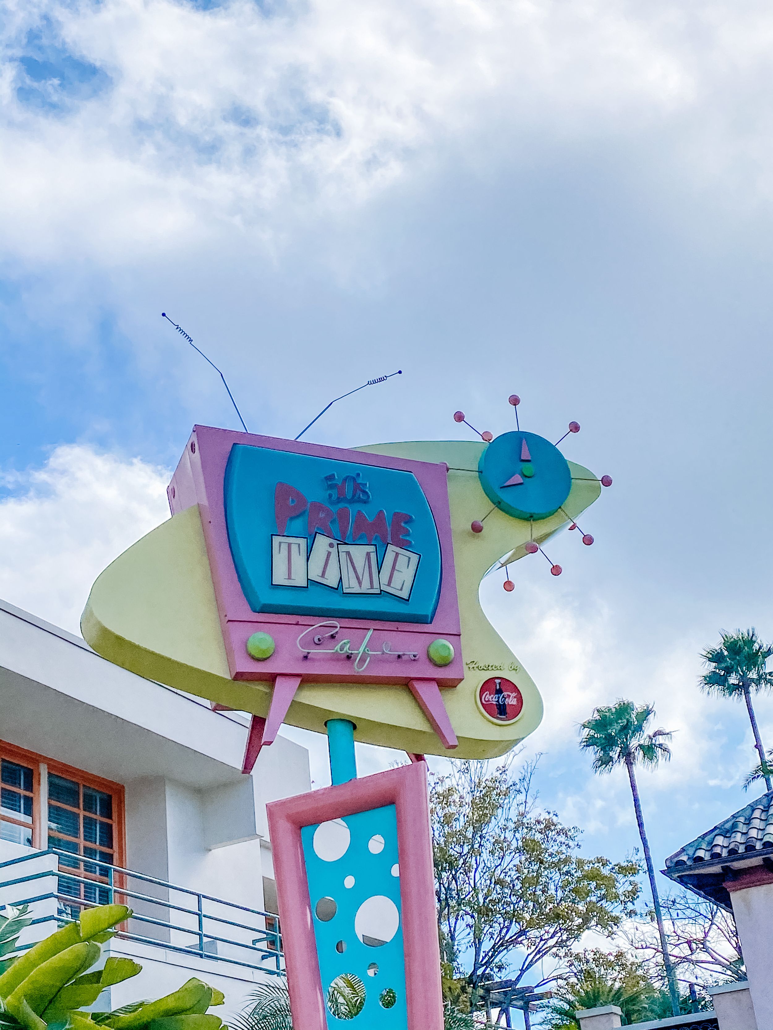 A retro-style pink and turquoise TV sign with vintage atomic design elements and palm trees against a blue sky.