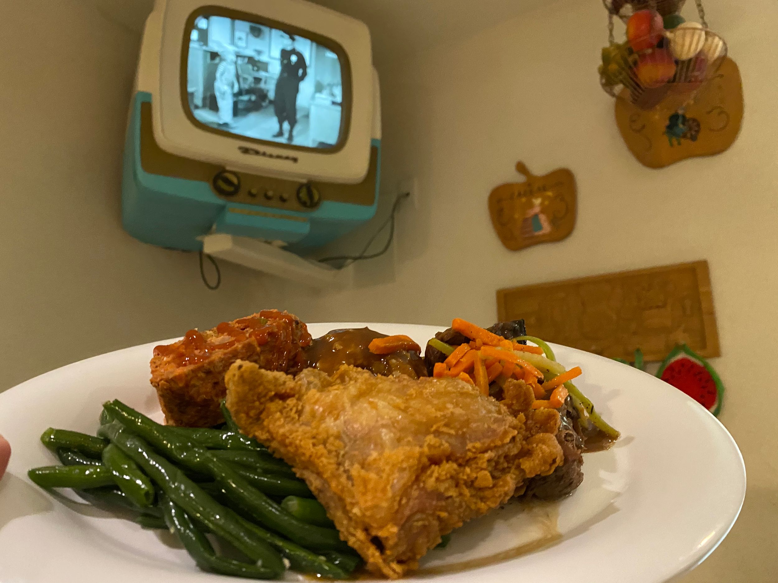 A plate of crispy fried chicken with green beans is served while a vintage turquoise television plays in the background.