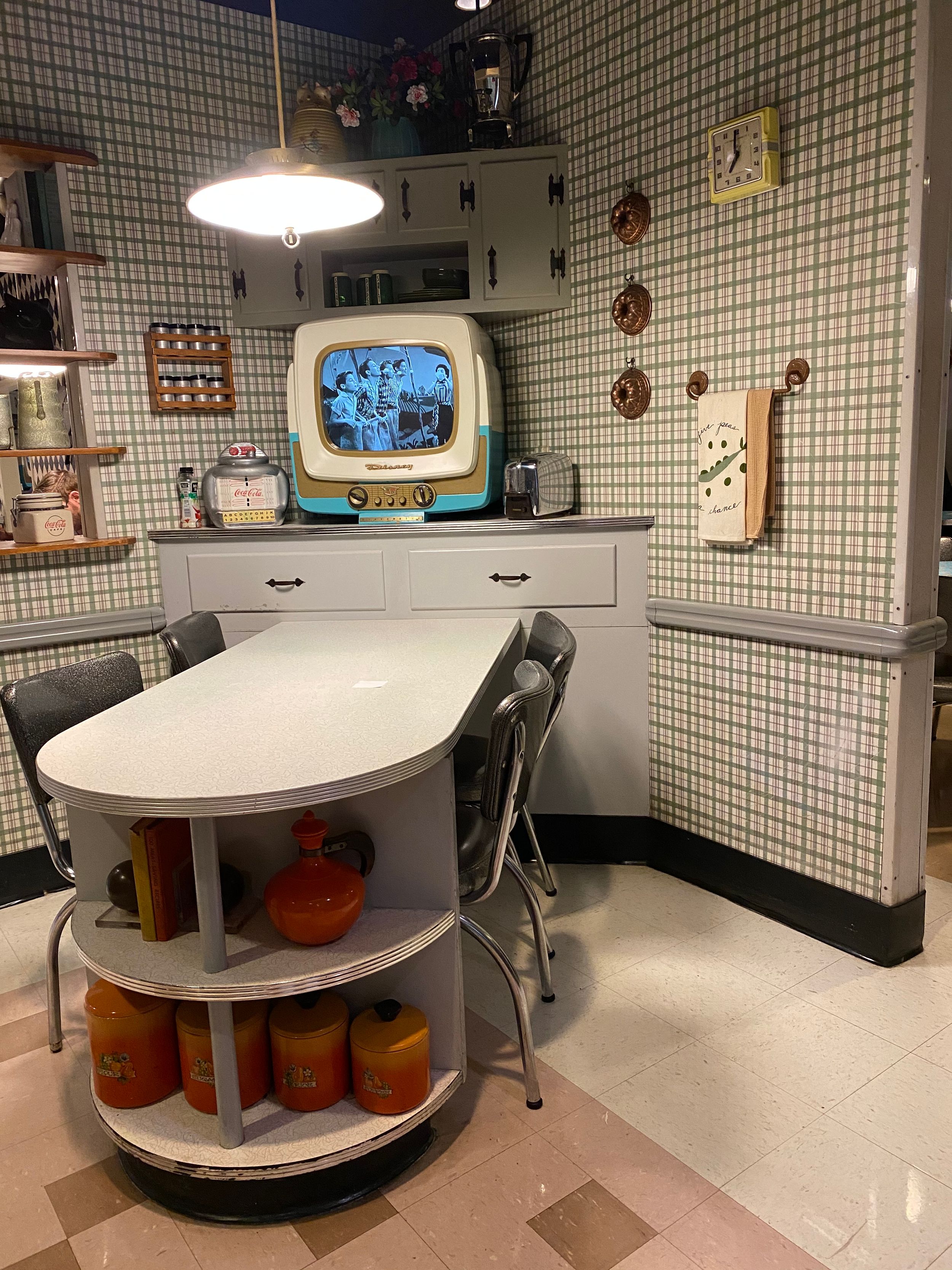 A retro 1950s kitchen corner with vintage TV, oval dining table, metal chairs, and orange storage containers below.