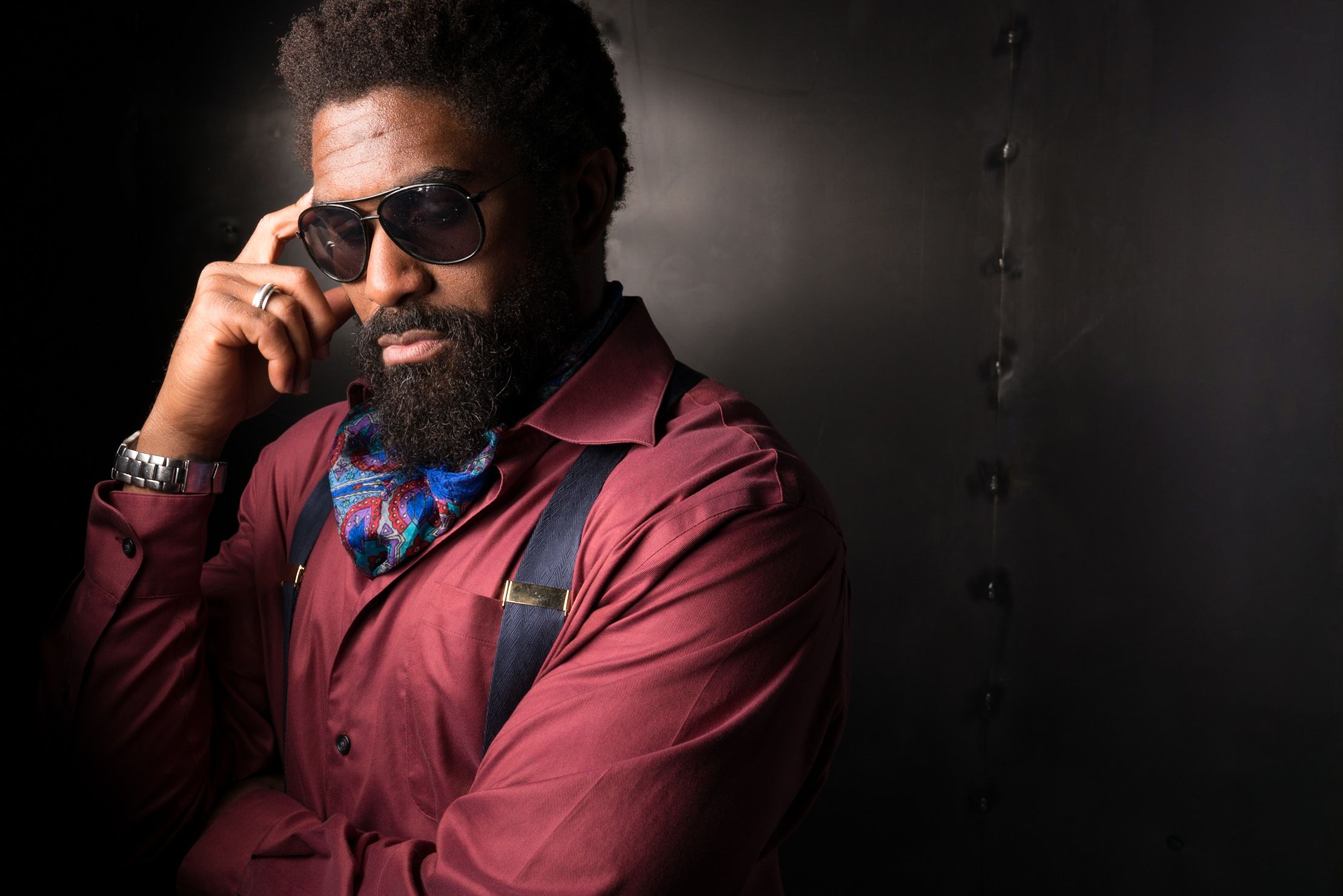 Stylish portrait of a man in sunglasses and burgundy jacket against a dark background.