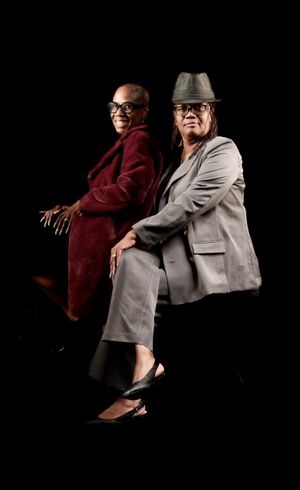 Two performers in formal attire pose back-to-back on a dark background, one in burgundy and one in gray with a hat.