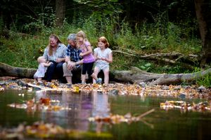 A family sitting together on fallen leaves by a reflective pond in an autumn forest setting.