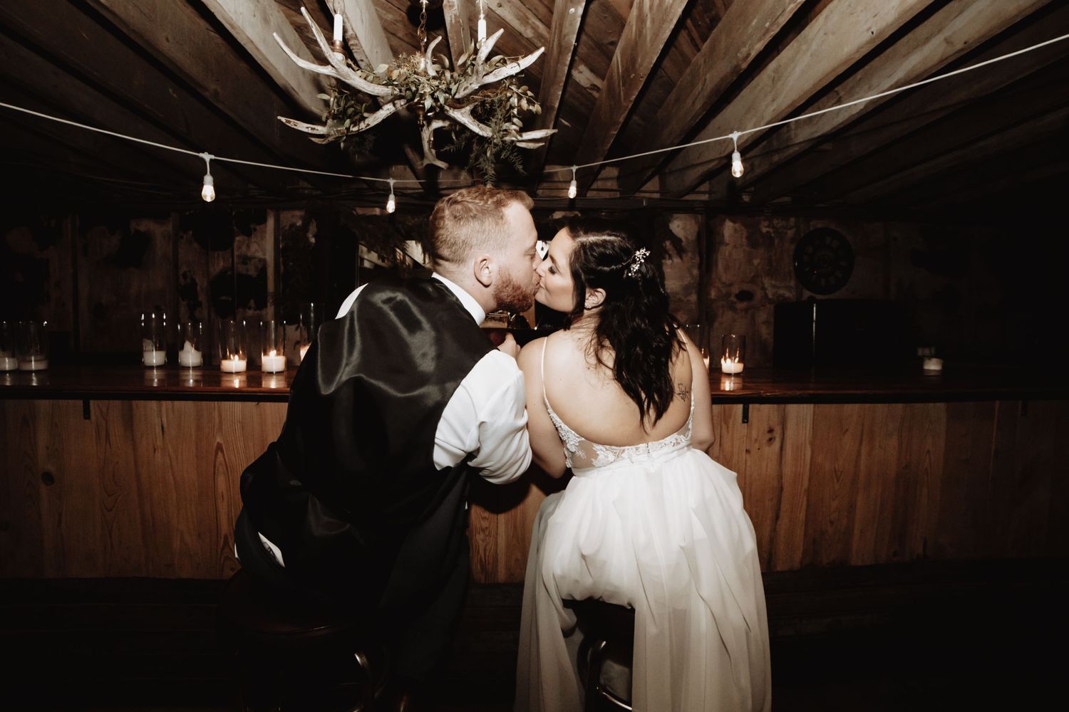 A romantic wedding couple sharing a kiss under rustic antler chandeliers in a barn reception venue.