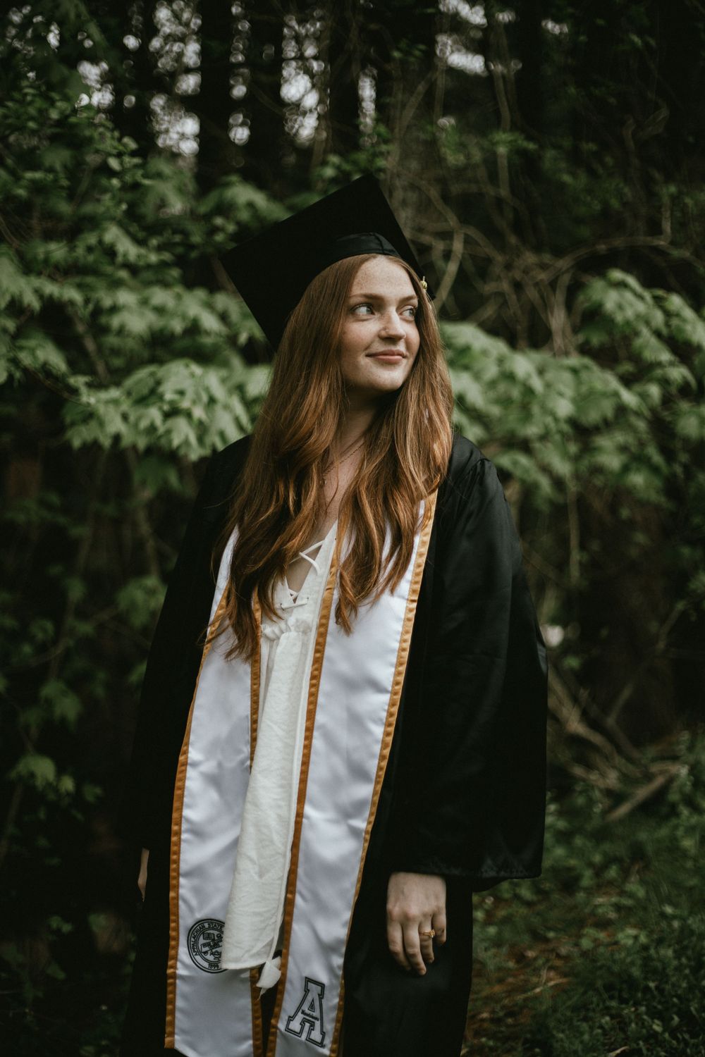 Graduate in cap and gown with gold stole stands in front of lush green forest setting.