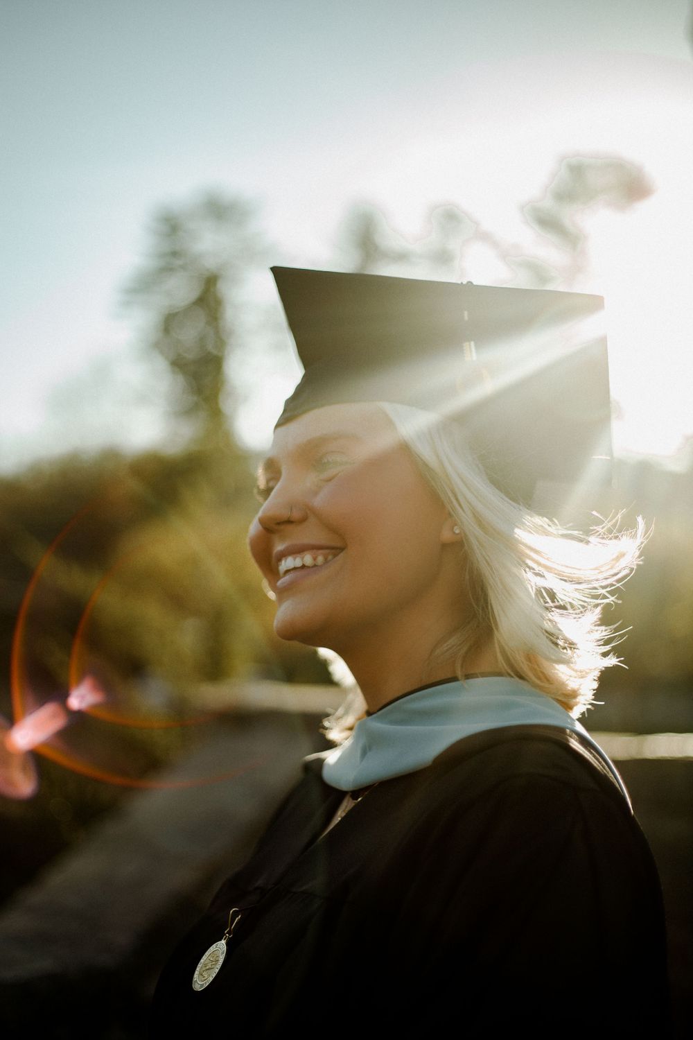 Graduate in cap and gown smiling in bright sunlight with lens flare effect.