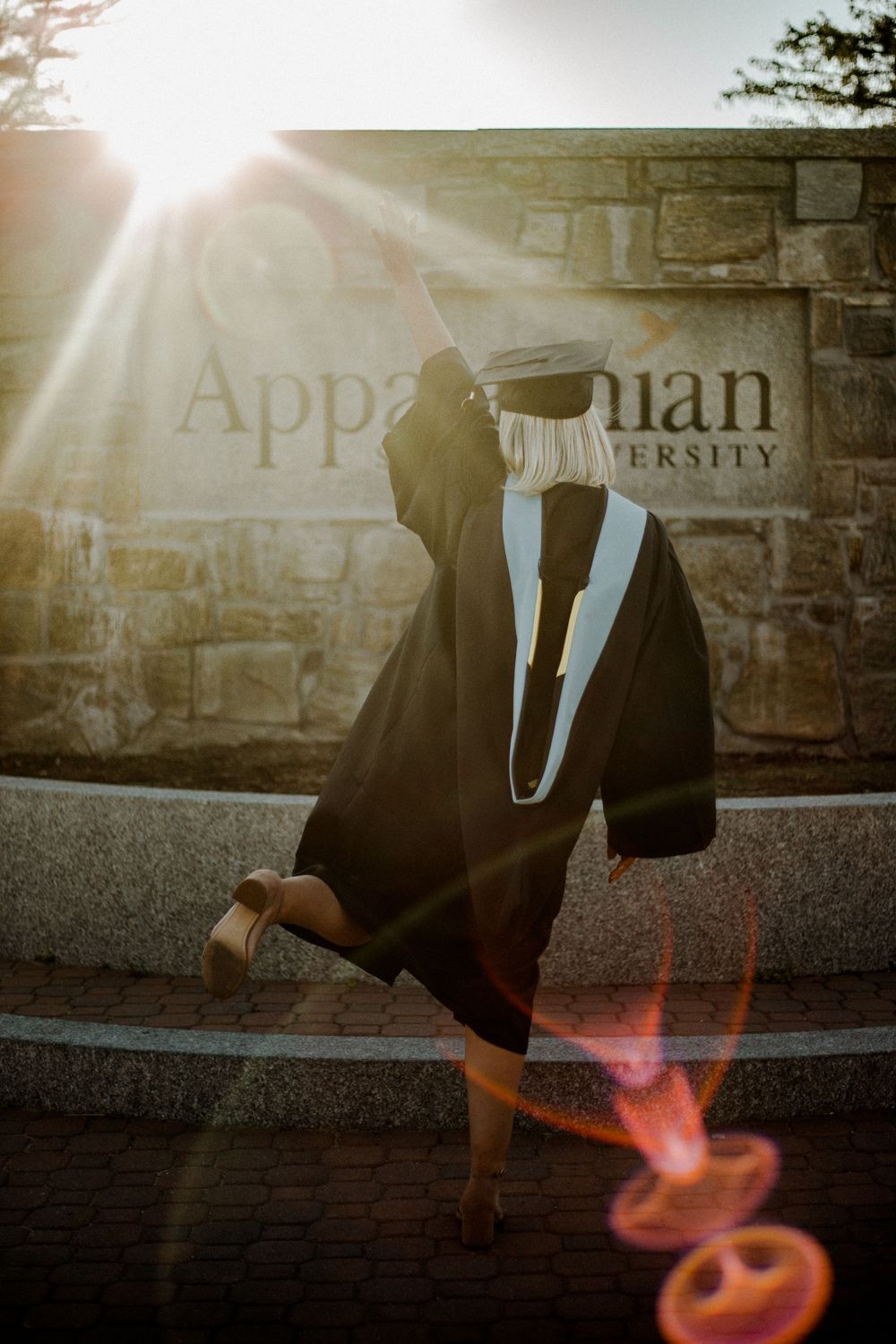 Graduation cap and gown silhouetted against stone wall with university signage at sunset.