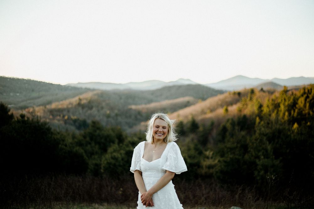 Woman wearing white dress stands in scenic mountain setting during golden hour.