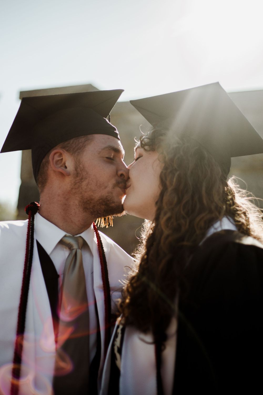 Graduates sharing romantic moment in caps and gowns with sun flare.