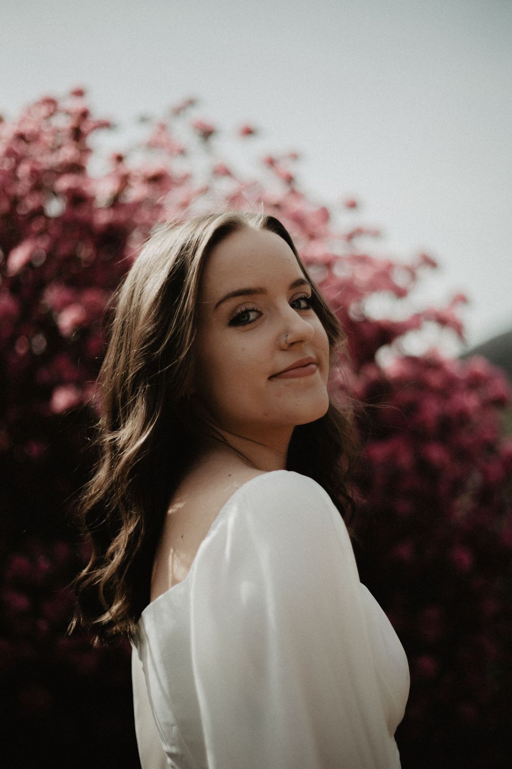 Portrait of woman in white dress against pink flowering trees in soft lighting.