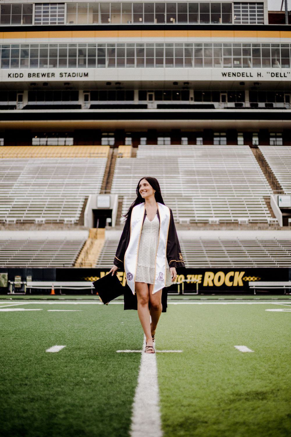 Graduate poses on football field center line at Kidd Brewer Stadium wearing white dress and cap.