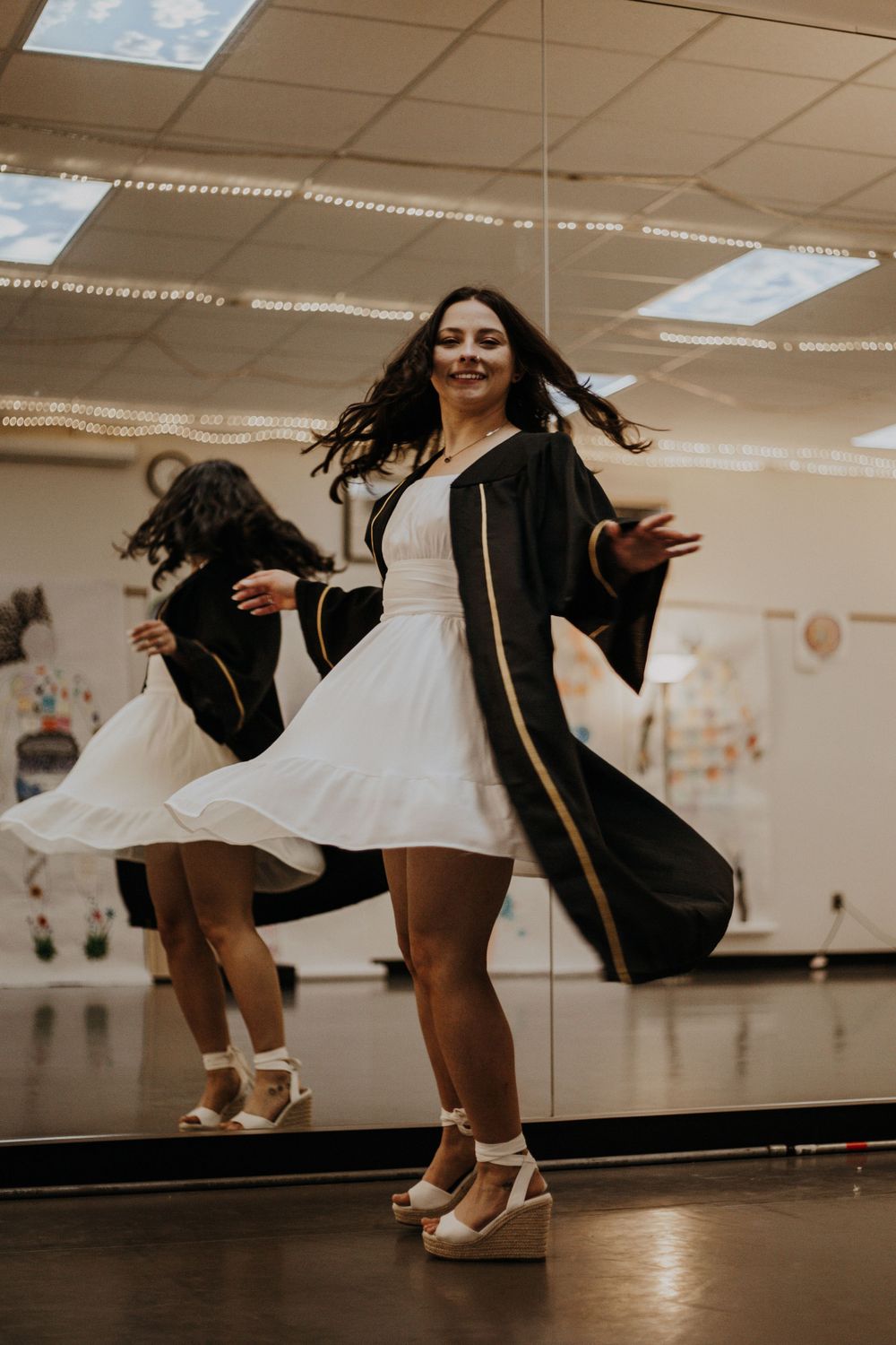 Graduate twirling in white dress and black graduation gown in classroom.