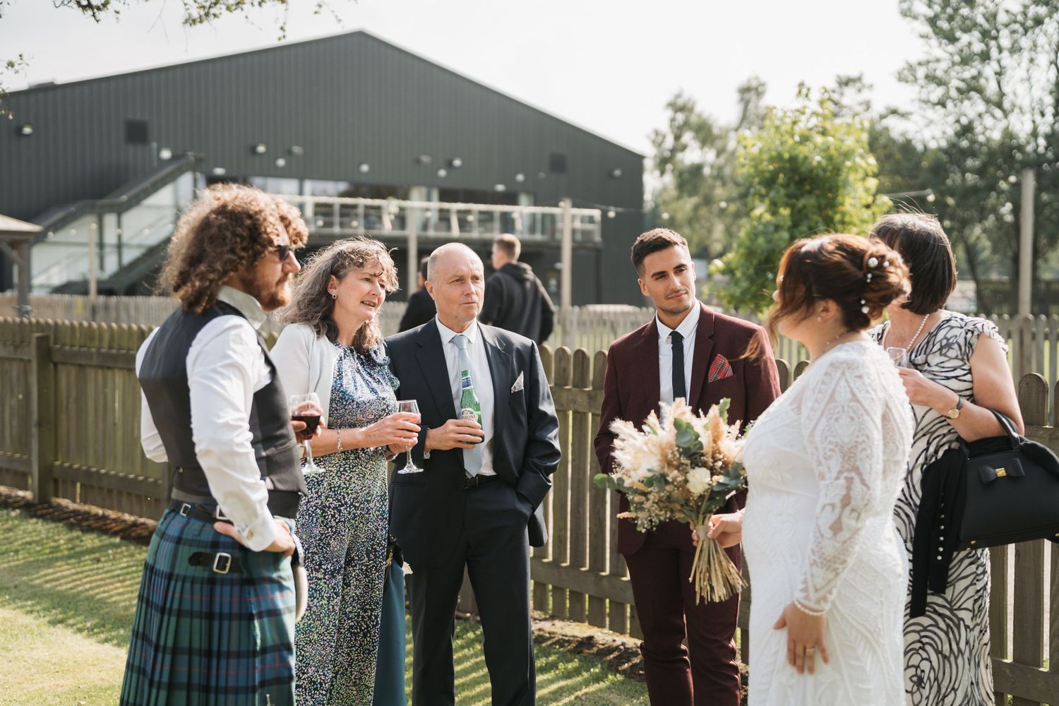 Wedding guests engage in cheerful conversation during an outdoor reception near a rustic building with a dark exterior.