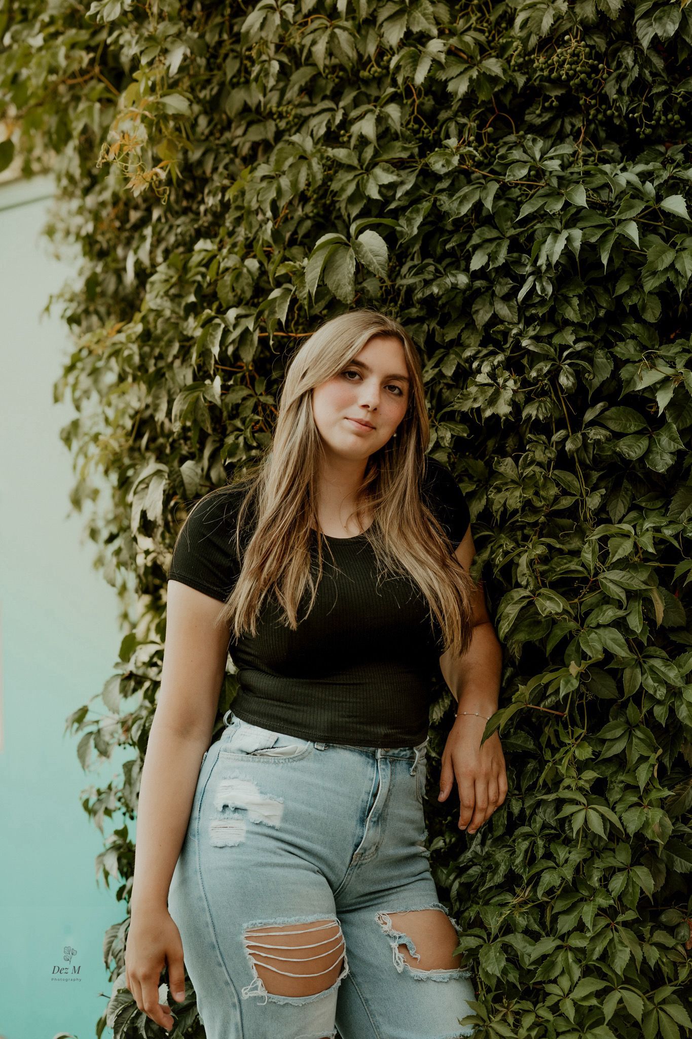 Person in black shirt and distressed jeans poses against ivy-covered wall in casual outdoor portrait.
