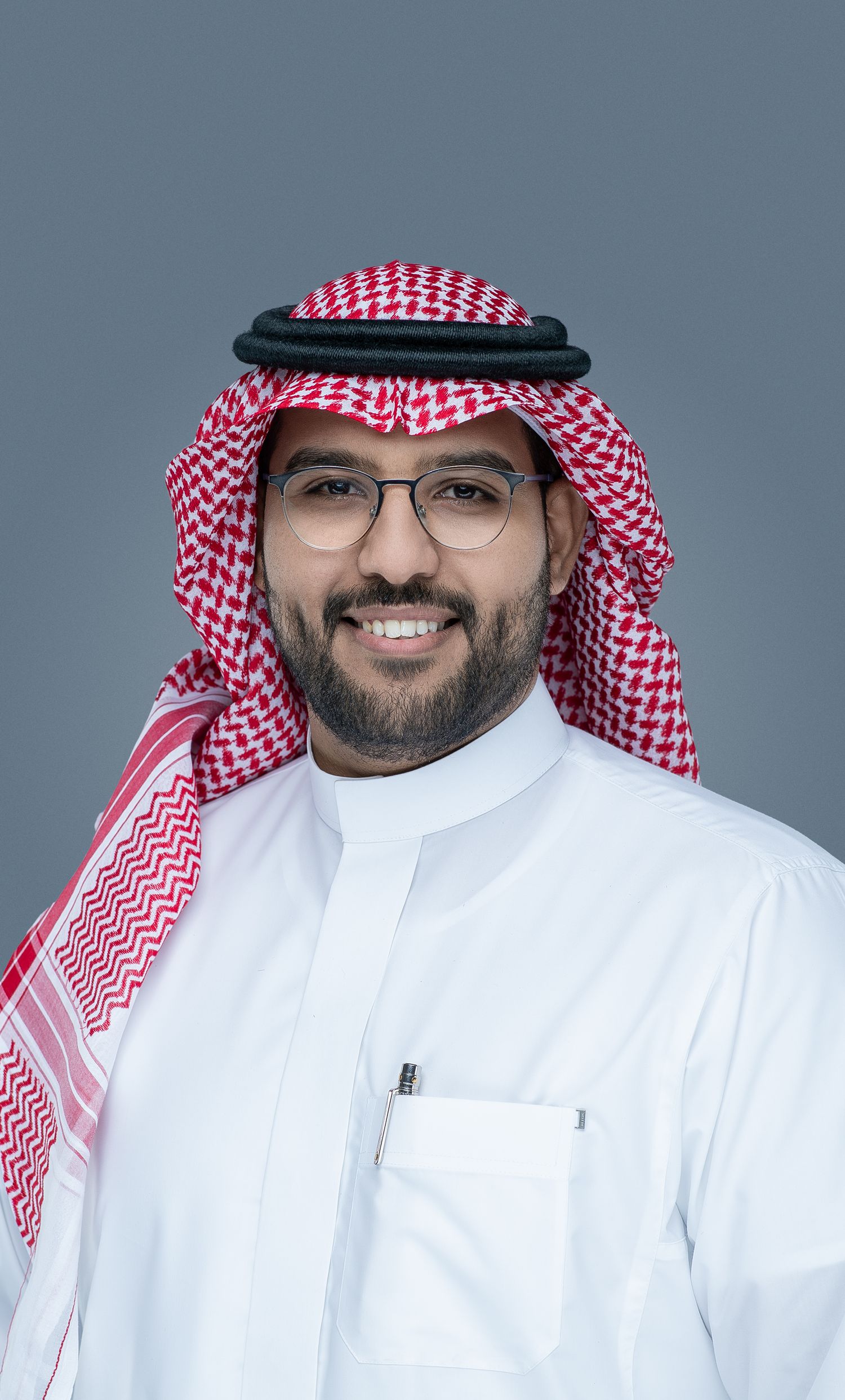 Portrait of person wearing traditional Saudi dress with white thobe and red and white checkered headdress against gray background.