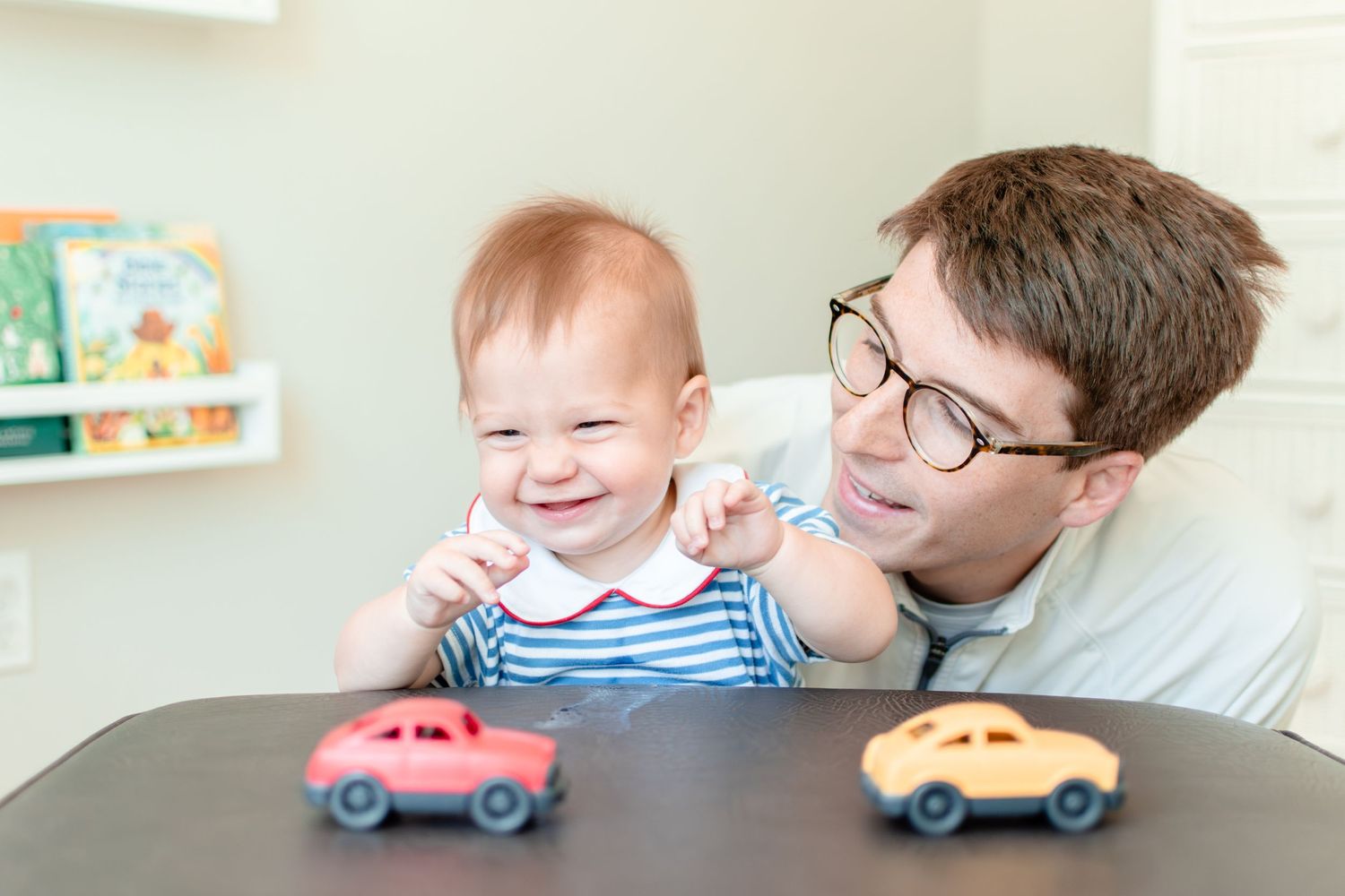 A baby and adult share a joyful moment playing with toy cars on a dark table against a light wall.