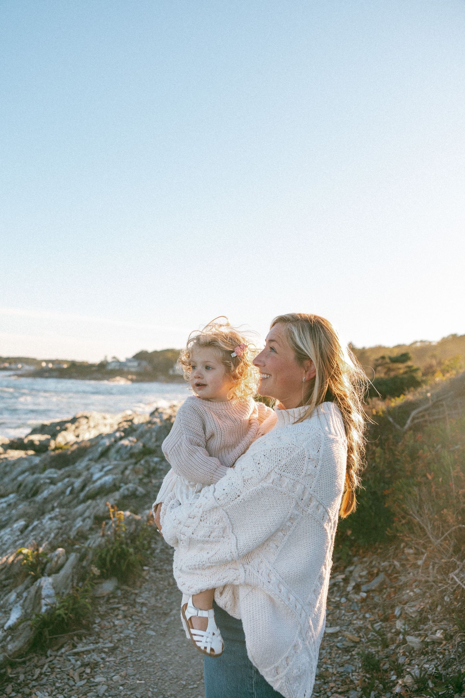 Two people embrace while standing on rocky coastal terrain at sunset with ocean views in the background.