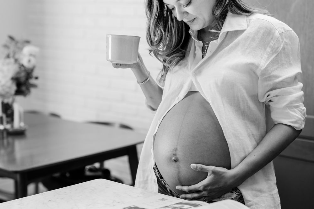 Tender maternity photo shoot showing pregnant woman in white shirt by window.