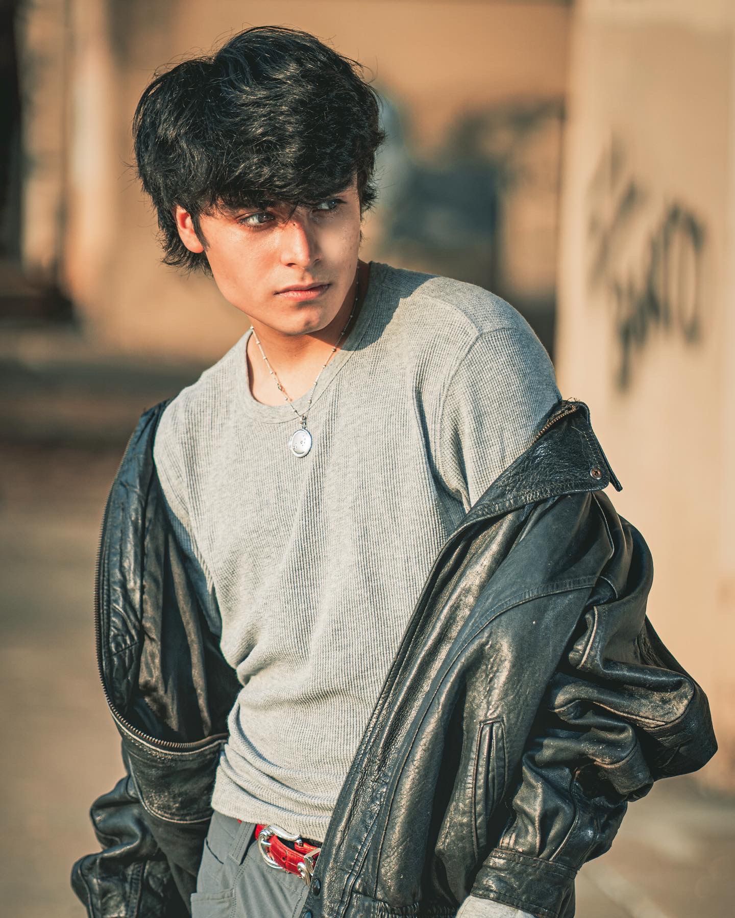 A figure wearing a leather jacket over gray henley poses casually against a blurred warm-toned background.