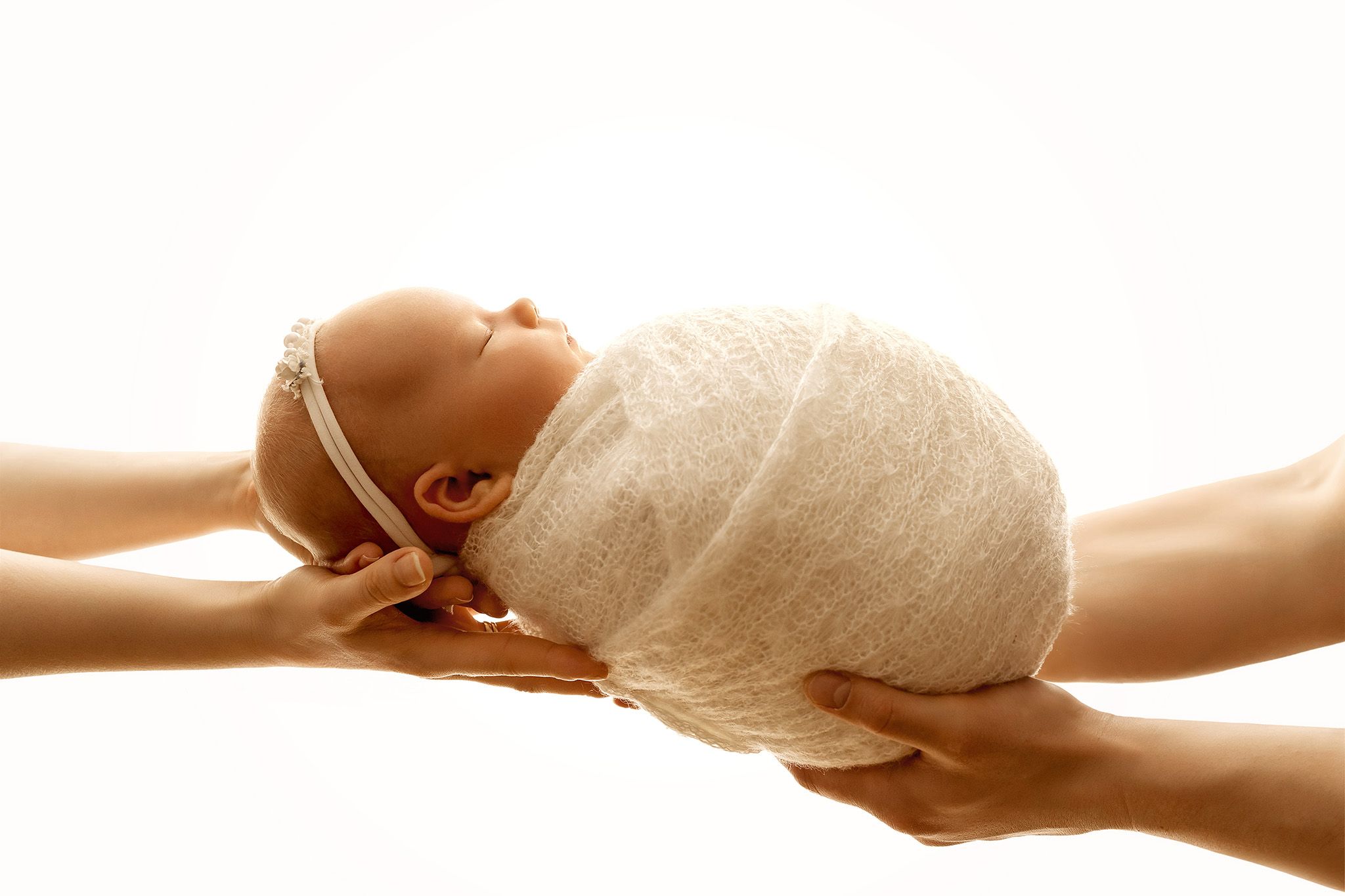 Newborn baby wrapped in a white lace wrap is hold by parents hand on white backdrop