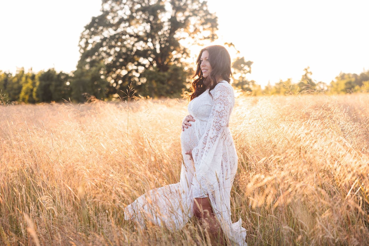 Pregnant woman in flowing white dress stands in golden wheat field at sunset while holding her belly.