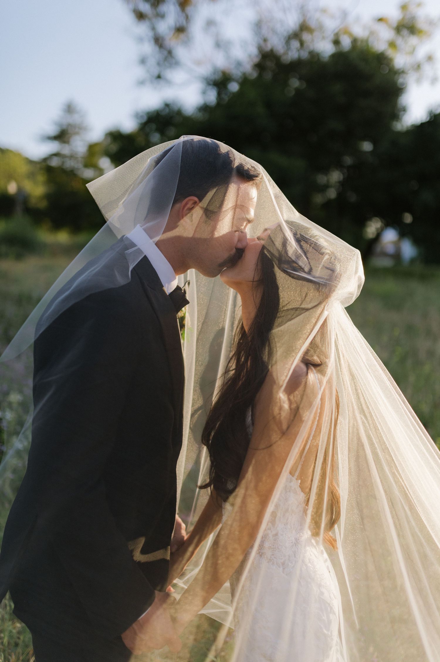 A romantic wedding moment captured as newlyweds share a kiss under a flowing veil in an outdoor garden setting.