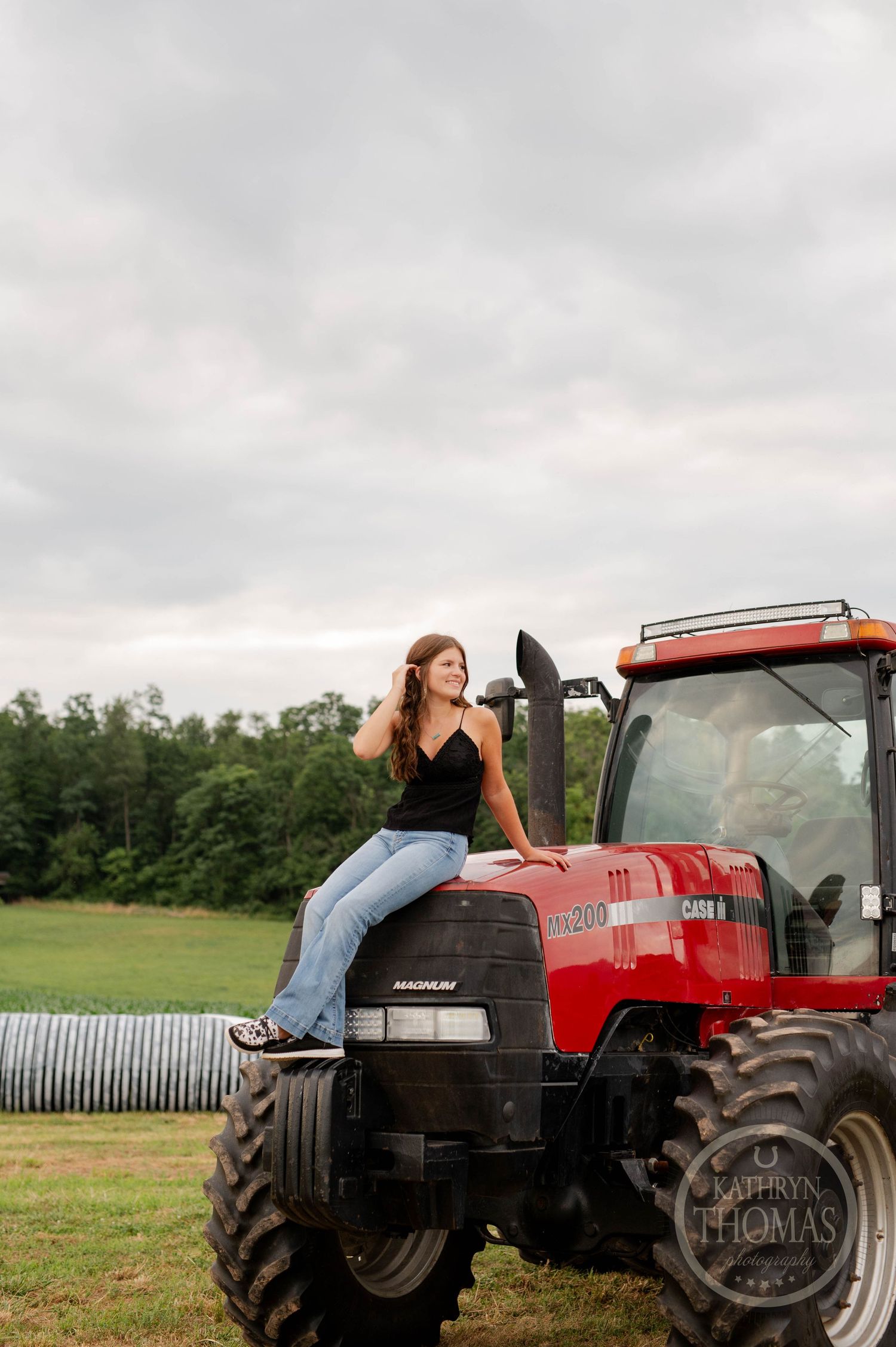 Woman in denim poses casually on hood of red Case tractor in rural setting.