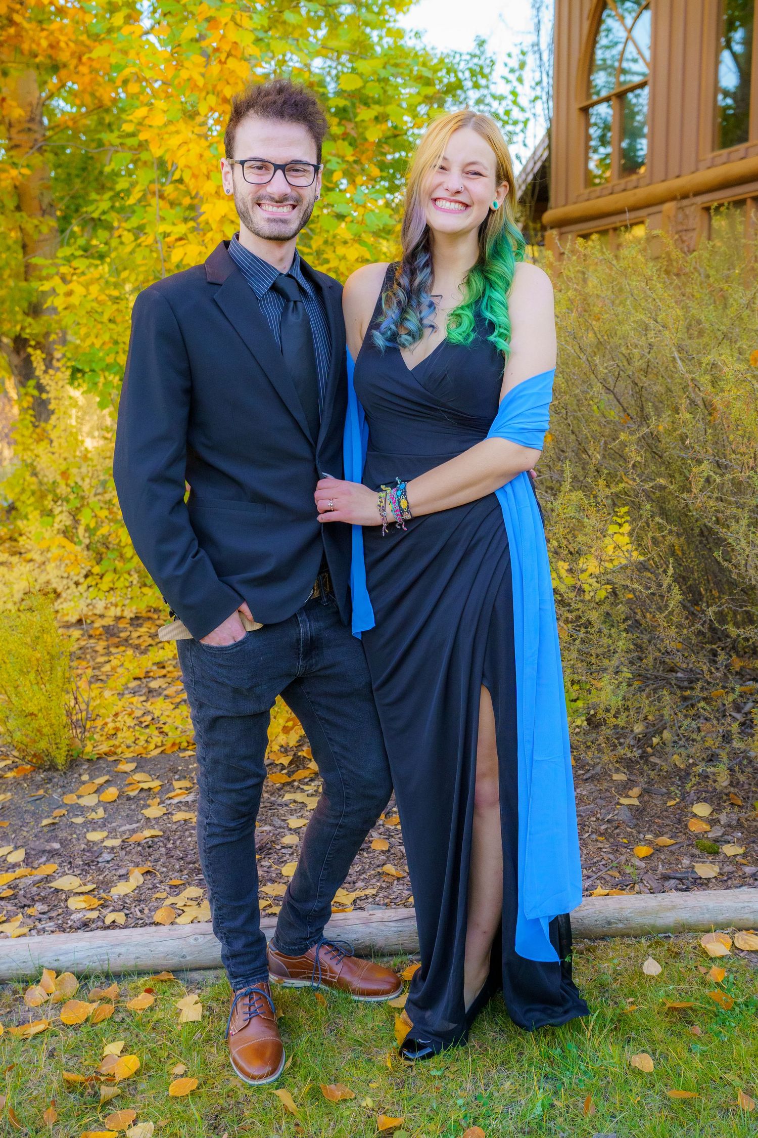 A couple poses outdoors in formal attire at a fall event, with vibrant autumn foliage in the background.