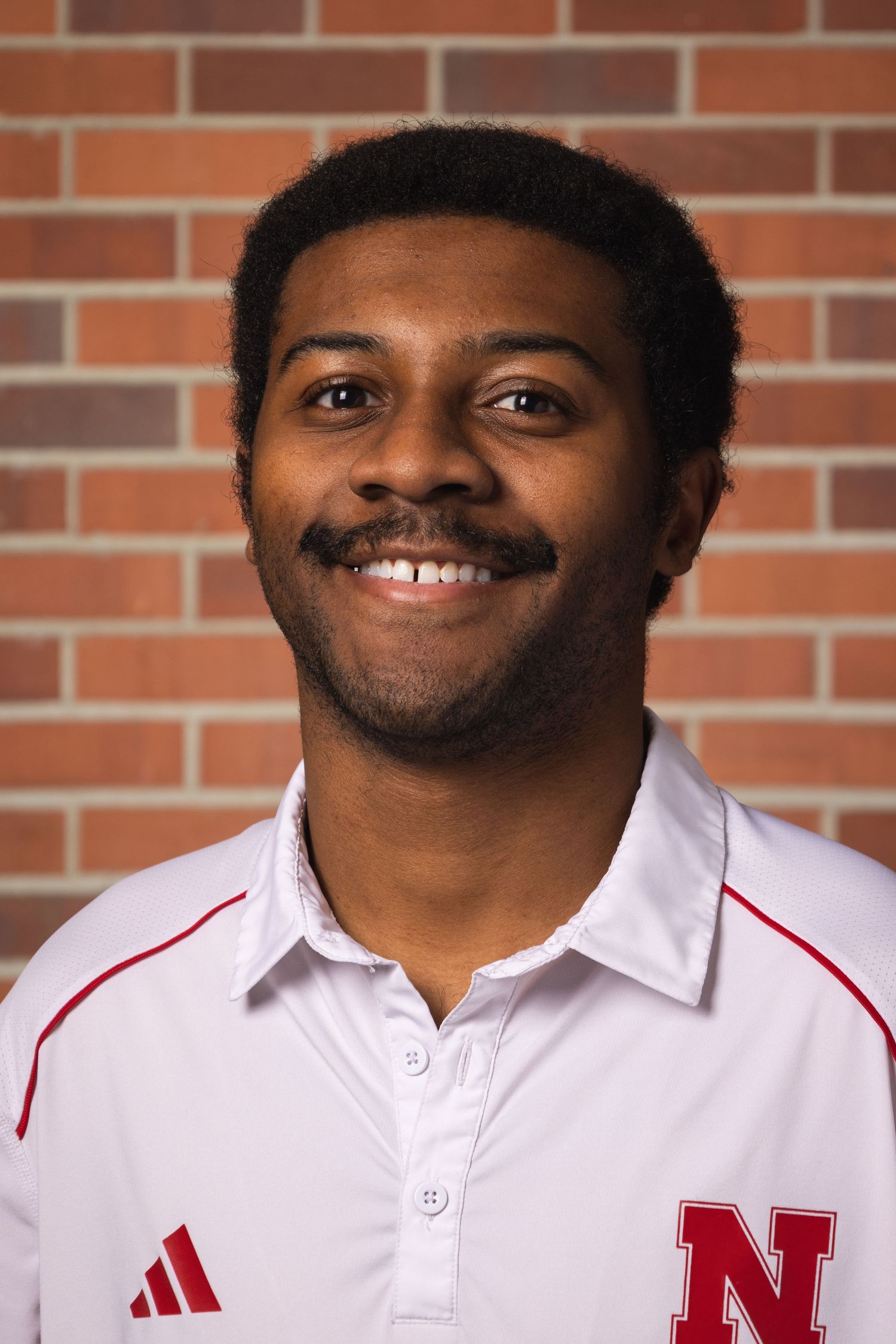 A person in a white Adidas polo shirt with red trim smiles against a brick wall background.