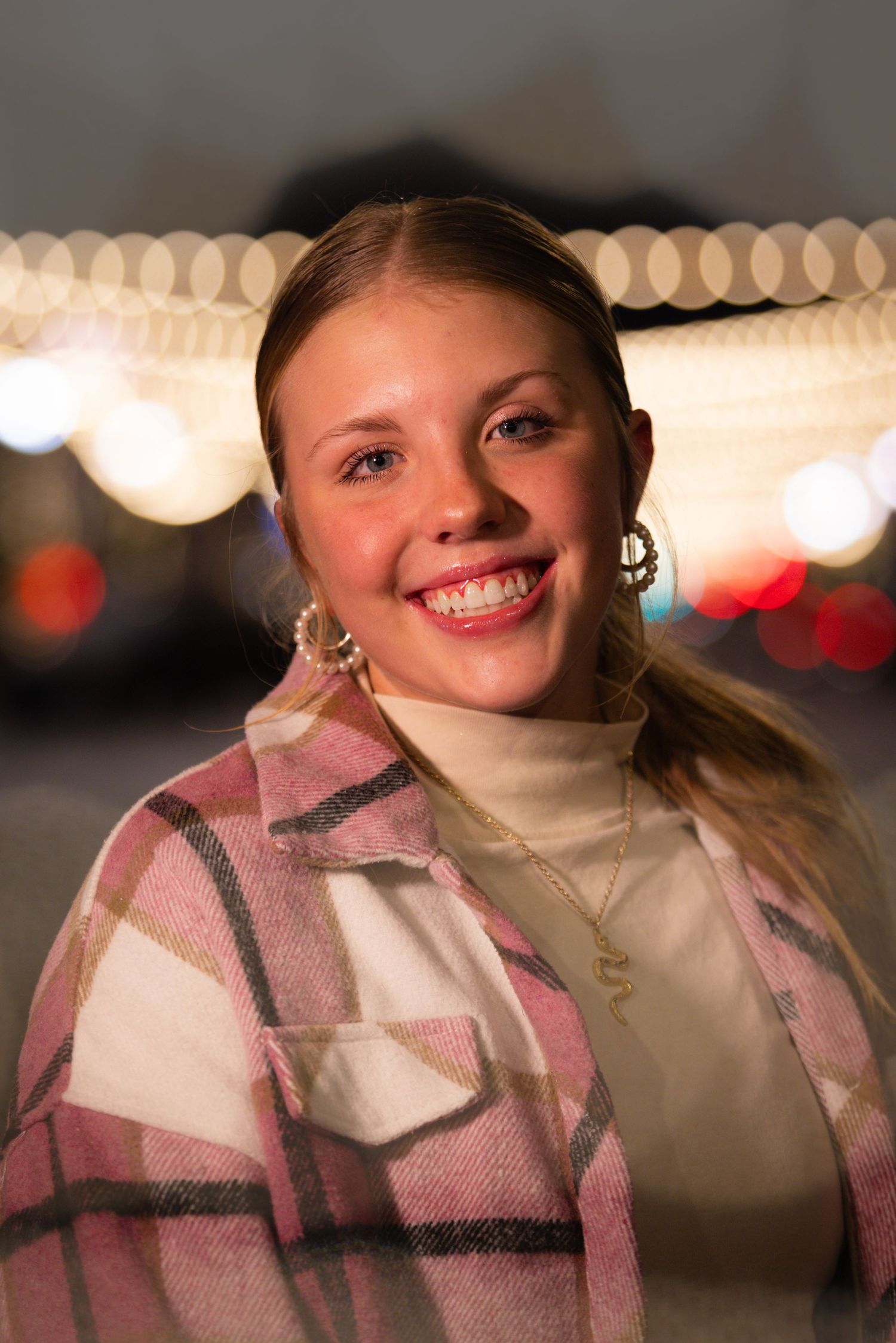 A person wearing a pink and white plaid scarf smiles warmly against a blurred background with bokeh lights.