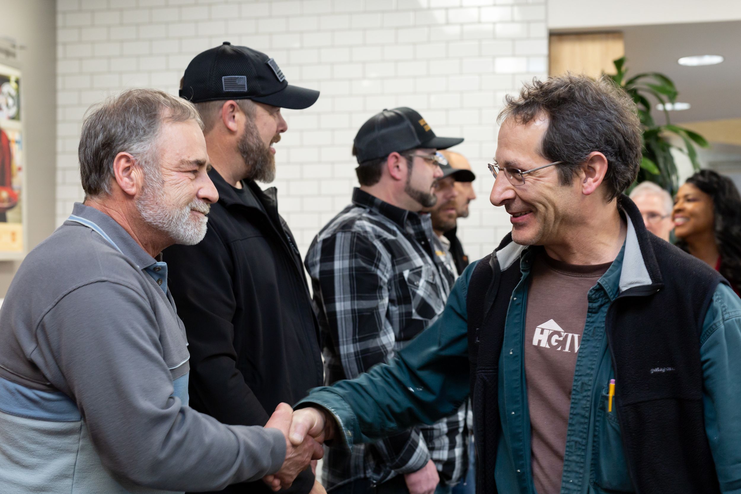 Two men shake hands warmly while sharing a laugh in an interior space with white tile walls.