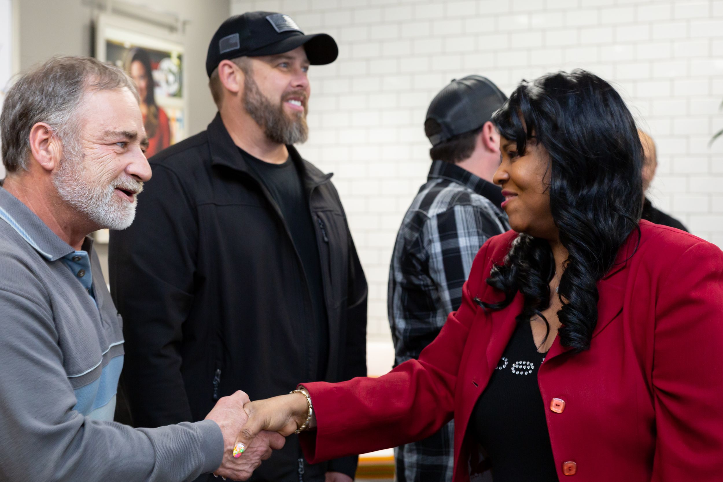 Two people shake hands warmly in a business setting while others look on in a white-walled room.